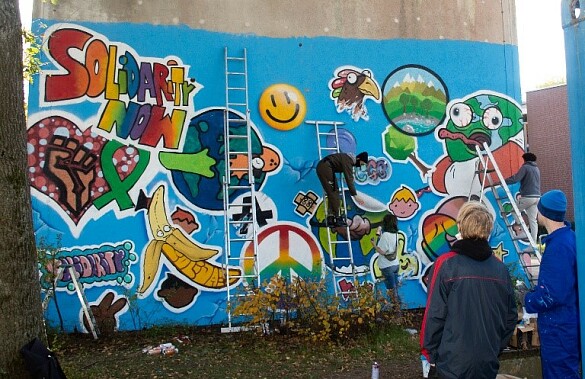 Ein Foto wo zwei Personen zwei Künstler:innen zu schauen, wie diese eines der Mottos der letzten Nacht der Jugend auf eine Wand sprühen.